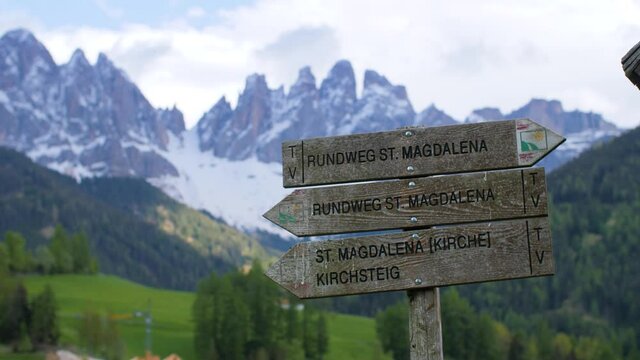 A signpost at the Santa Maddalena hiking trail in the Italian alps, Dolomites. The sign indicates different roads in the beautiful Val di Funes valley with view of the Odle mountain range, Italy. 4K.