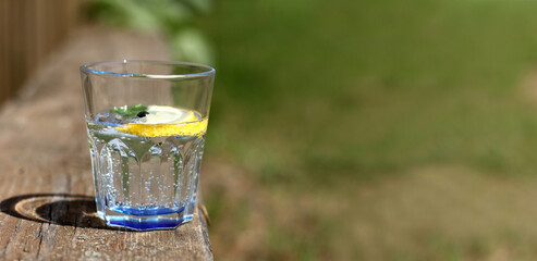 glass of soda water decorated with lemon, mint leaf and currant berry on a bench on a sunny summer day. cool refreshing natural drink