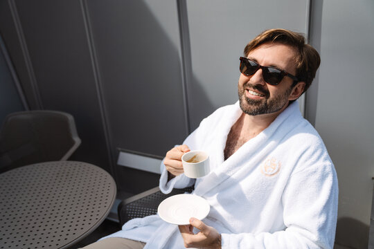 Smiling mid aged bearded man in bathrobe drinking coffee in the morning sitting on a balcony