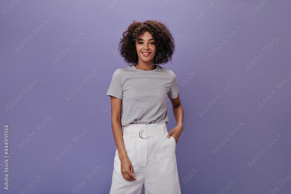 Sticker Snapshot of curly woman in white shorts against purple background. Charming brunette girl in grey tee smiling on isolated backdrop