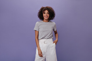 Snapshot of curly woman in white shorts against purple background. Charming brunette girl in grey tee smiling on isolated backdrop