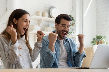 Overjoyed mixed race couple sit in kitchen looking at laptop screen celebrate online lottery...