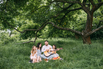 Cheerful caucasian family sitting on soft blanket at garden and singing songs, having fun. Handsome father playing guitar for his charming wife and two cute daughters.