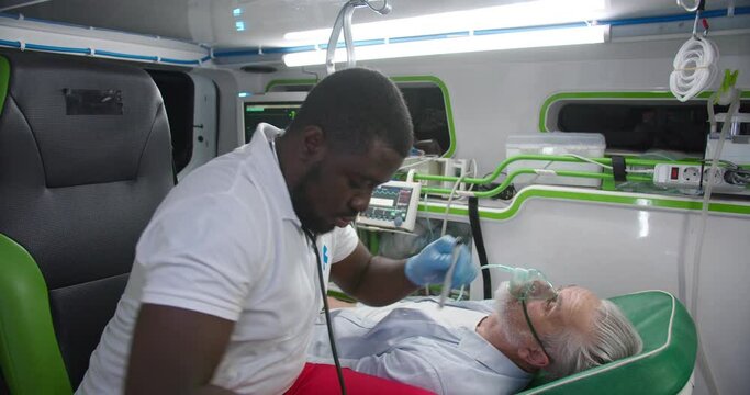African American paramedic saving life of old man in ambulance. Medic giving oxygen to patient and checking eyes reaction. Reanimation. Rescuing paramedical work. First aid of doctor. Emergency.