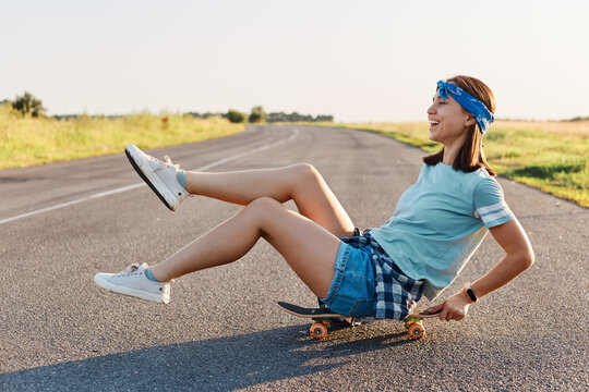Side view of happy emotional woman sitting on skateboard on the street with raised legs, overjoyed female wearing casual clothing having fun outdoors, positive lifestyle.