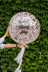 There's a wall of ivy. In front of it, a transparent balloon containing pink confetti is floating in the air and a happy birthday message is written.