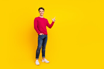 Full size photo of optimistic brunet millennial guy point empty space wear red sweater jeans isolated on yellow color background