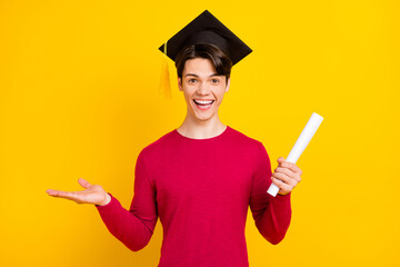 Photo of happy pretty guy dressed red sweater bachelor headwear holding diploma arm empty space isolated yellow color background