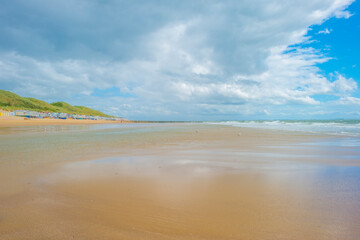 Sunlit waves on the yellow sand of a sunny beach along the North Sea illuminated by the light of a colorful sun and a blue cloudy sky in summer, Walcheren, Zeeland, the Netherlands, July, 2021
