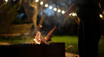 Wooden logs burning in the grill in the night garden decorated with lanterns. Gloved hand lays logs...