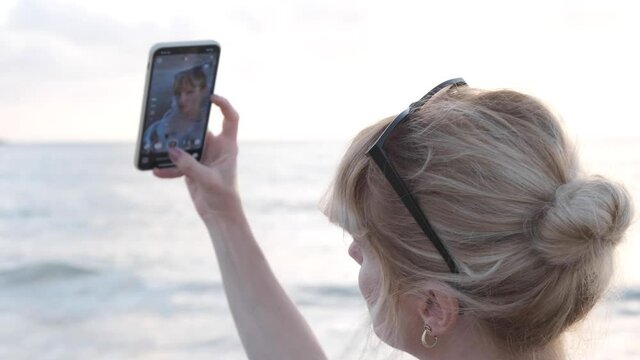 young woman taking selfie on the beach. beautiful blond woman on a holiday on the shore taking pics with her smartphone. posing to the camera creating reels for social media