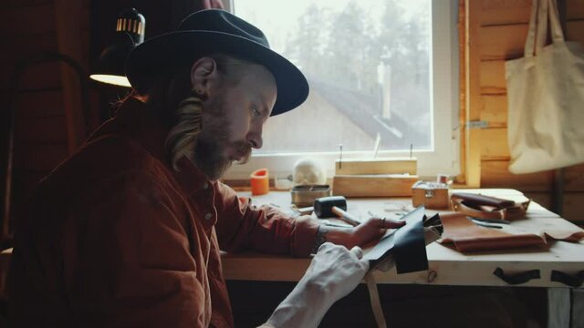 Zoom in shot of bearded craftsman in fedora hat sitting at desk in workshop and working on leather craft