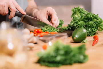 The man is in the kitchen. Male hands hold a knife and cut vegetables and green salad.