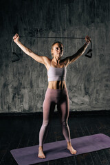 Muscular young athletic woman with perfect beautiful body wearing sportswear holding resistance band above head standing on mat. Caucasian fitness female training with stretching expander in studio.
