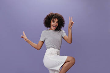 Lady in bright outfit jumps on purple background and shows peace sign. Attractive woman in white shorts and gray tee dancing