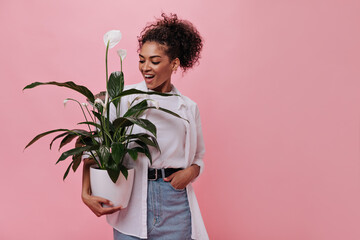 Woman in bright outfit holds plant on pink background. Happy curly girl in white tee and skirt...
