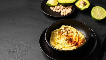 Black ceramic bowl with chickpea hummus on black background with chickpea beans , avocado, smoked paprika and lime multigrain bread,