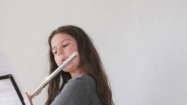 Little girl looking at the notes and playing the flute in bright room. Children musicians concept. Out-of-school activities, home distance learning.