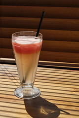 Fresh grapefruit juice in glass with black straw on wooden background. Light and shadow from the window. Thin lines of light on the wall and table. Shadow from the blinds.
