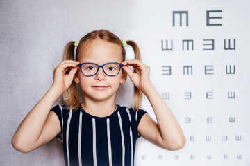 Happy little girl wearing glasses taking eyesight test before school with blurry eye chart at the background, child's vision examination - obrazy, fototapety, plakaty