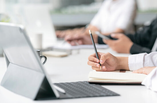 Cropped shot of woman hand writing on notebook with pencil while sitting with her teamwork at office.