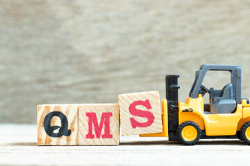 Toy forklift hold letter block S to complete word QMS (abbreviation of quality management system) on wood background