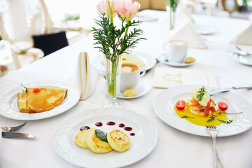 Three cheesecakes and pancakes on a white plate with jam, background of a vase with flowers, restaurant