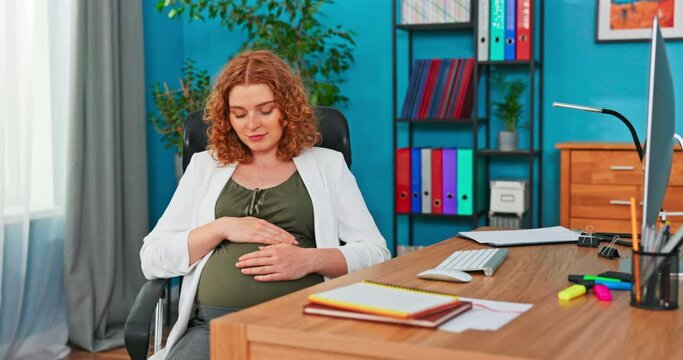 The head of company sits on a swivel chair behind a desk in company's office, woman strokes, touches and looks at her pregnant belly, she smiles, happy, not resting just working, no maternity leave