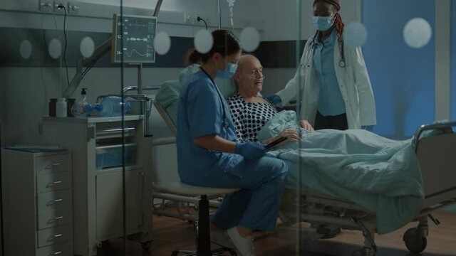 Nurse and african american medic checking on patient in hospital ward bed with help of modern technology. Sick elder man with nasal oxygen tube and oximeter laying in bed for treatment