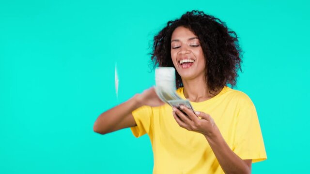 African woman with happy face scatters money. Lady is overspend USD currency. Girl is flush with dollars on teal studio background.