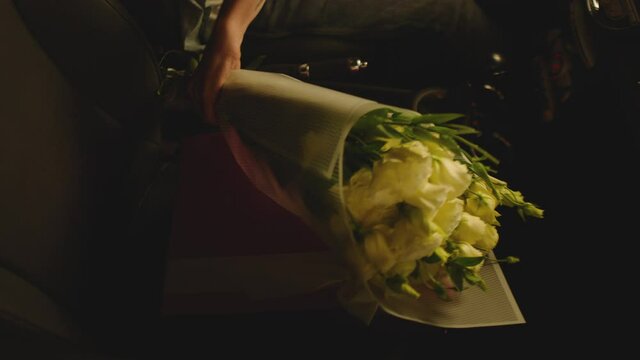 Man Putting Down White Flowers And Purple Gift Box Inside Car . Male's Hand Taking Beautiful Roses Or Tulips And Surprise Wrapped Present From Front Seat Of Car .  Black Car Interior . Slow Motion 