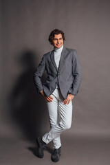 Tall handsome man dressed in white turtleneck, white jeans and grey jacket posing on the grey background