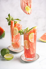 Two glasses with tall long drink with watermelon, lime slices - and mint - a hand pressing lime juice
