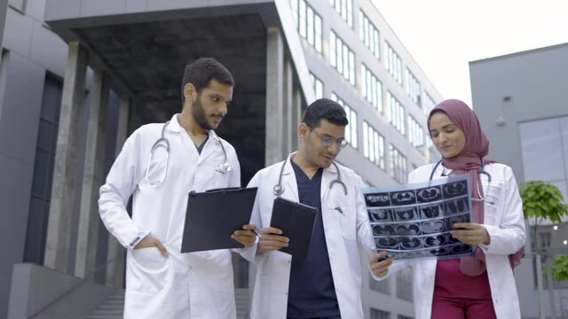 Three doctors, woman in hijab and two men in medical apparel, discussing patient's x-ray tomography scan, walking outside on the background of modern hospital with stairs