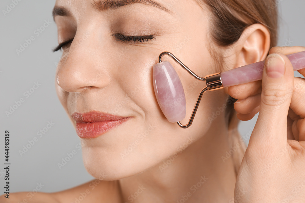 Sticker Young woman using natural rose quartz face roller on light grey background, closeup