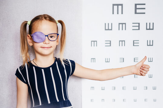 Little girl wearing glasse and eye patch or occluder with her thumb up and blurry eye chart at the background, amblyopia (lazy eye) treatment