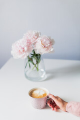 Female hand with cup of coffee and beautiful light pink peonies flowers in a vase on white table. Cozy breakfast, morning coffee. Enjoy the moment, take a break. Minimalist. Vertical card. Copy space