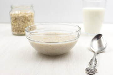 Oatmeal porridge cooked with milk, a glass of milk, a spoon, a plate on a white wooden table