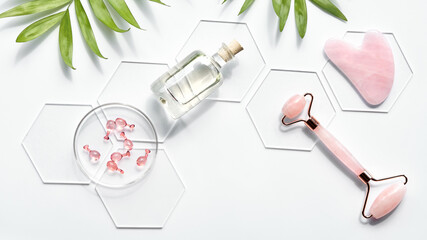 Pink Quarts stone face roller, serum capsules in round glass dish, hexagons, palm leaves. Gua sha stone for beauty facial massage therapy. Natural cosmetics laboratory. Panoramic flat lay, copy-space.