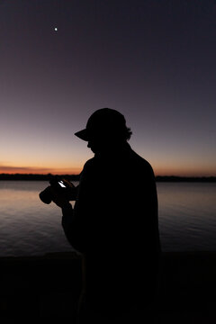 Un fot&oacute;grafo hombre con gorra a un costado de un rio revisa su c&aacute;mara en el atardecer