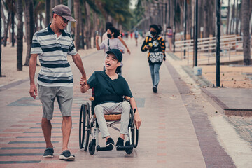 Confident disabled teen boy on wheelchair and father on the street in front of the beach, Lifestyle of disability kid activity in the travel holidays with family, Candids in public places concept.