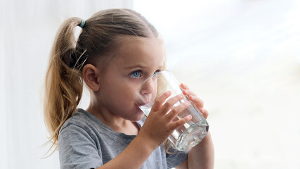 Little blonde girl in white dress drinks water from a glass indoors. Cute child is drinking a cup of water - obrazy, fototapety, plakaty