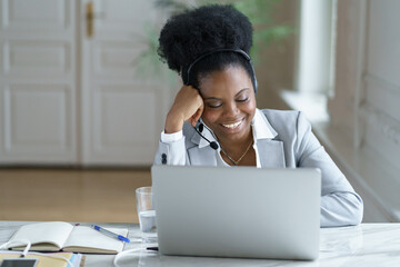 Friendly customer support operator in earphones work on laptop computer. African american female...