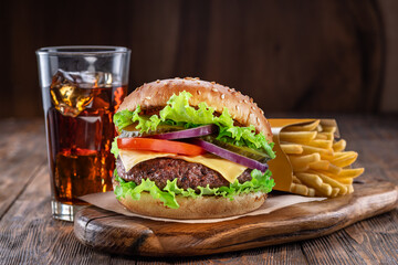 Delicious burger with cola and potato fries on a wooden table with a dark brown background behind. Fast food concept. - obrazy, fototapety, plakaty