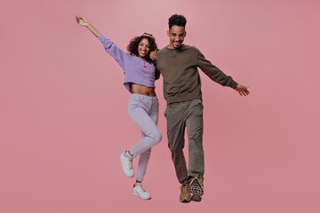 Curly woman and man in street style outfit posing on pink background. Cheerful guy in brown jeans and shirt hugs his girlfriend on isolated