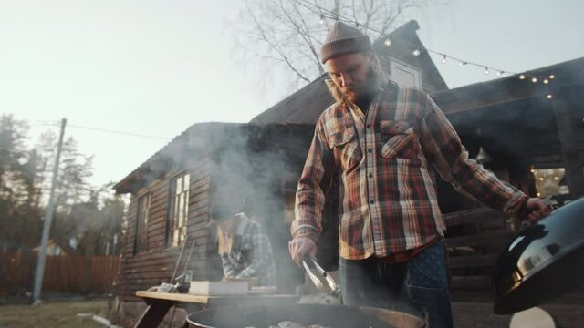 Low angle shot of man taking off lid frim bbq grill and using tongues for turning meat while cooking food outdoors in backyard