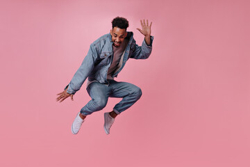 Active guy in denim outfit jumping on pink background. Dark-skinned brunette man in jeans moves on isolated backdrop