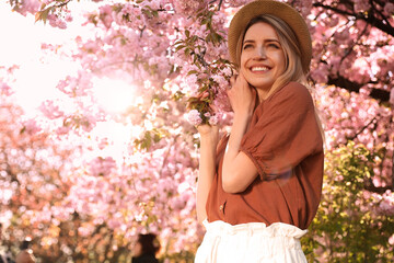 Young woman wearing stylish outfit near blossoming sakura in park. Fashionable spring look - obrazy, fototapety, plakaty