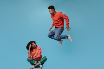 Obraz premium Happy girl in orange sweatshirt sitting. Man jumping on blue background. Portrait of guy in jeans moving and girl chilling on isolated