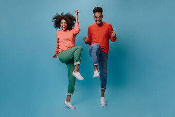 Fototapeta na wymiar Shouting girl and man in stylish outfits jumping on blue background. Emotional guy and brunette woman in jeans dance and move on isolated